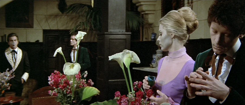 Whity (1971) movie still: medium — A dimly lit room features dark wood paneling and decorative floral arrangements on tables. A woman in a pink…