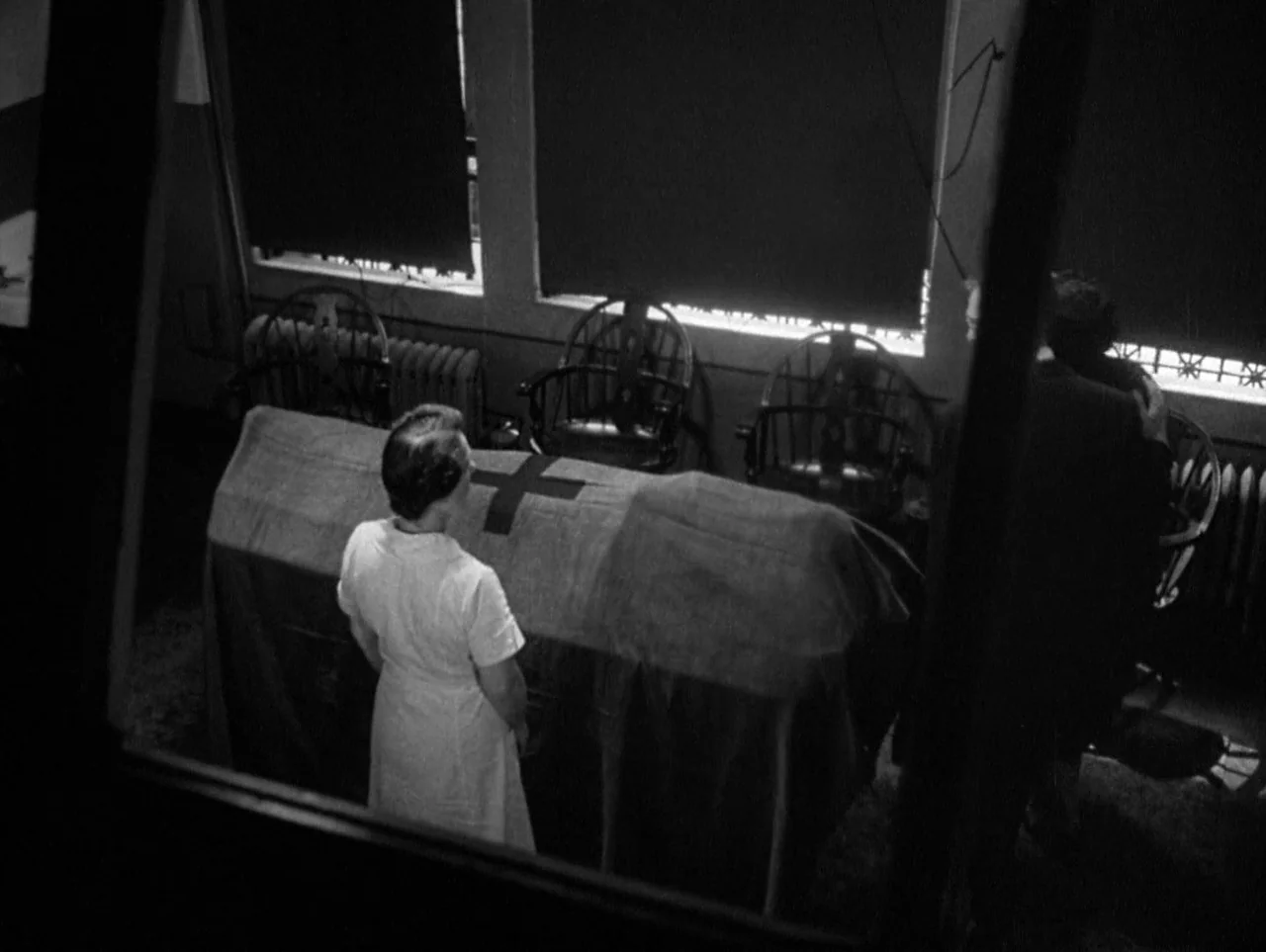 The Naked City (1948) movie still: wide — An interior chapel space features windows with dark shades and wooden chairs. A nurse and a man in a suit…