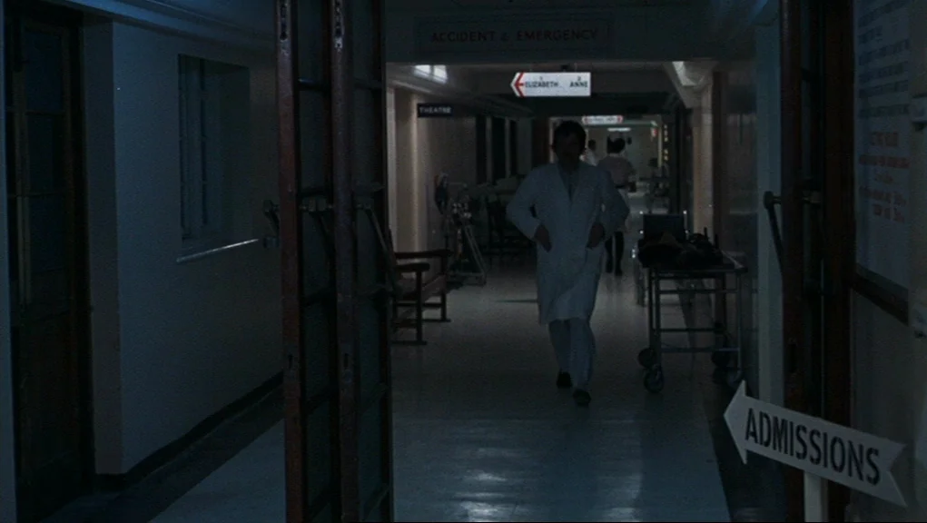 Frenzy (1972) movie still: medium — A dimly lit hospital corridor with overhead directional signage for emergency departments. A male doctor in a…