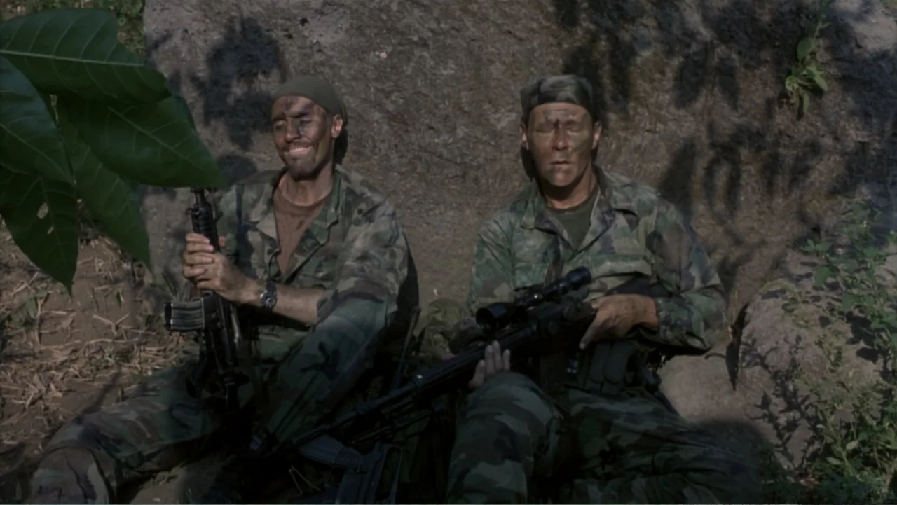 Behind Enemy Lines (2001) movie still: medium — A dense outdoor location featuring two soldiers resting against a large gray rock face. The soldiers…