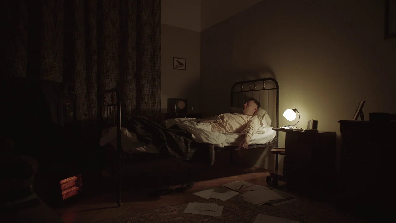 The Zone Of Interest (2023) movie still: wide — A dimly lit room features a metal frame bed and a cluttered floor with papers. A man sleeps in the…