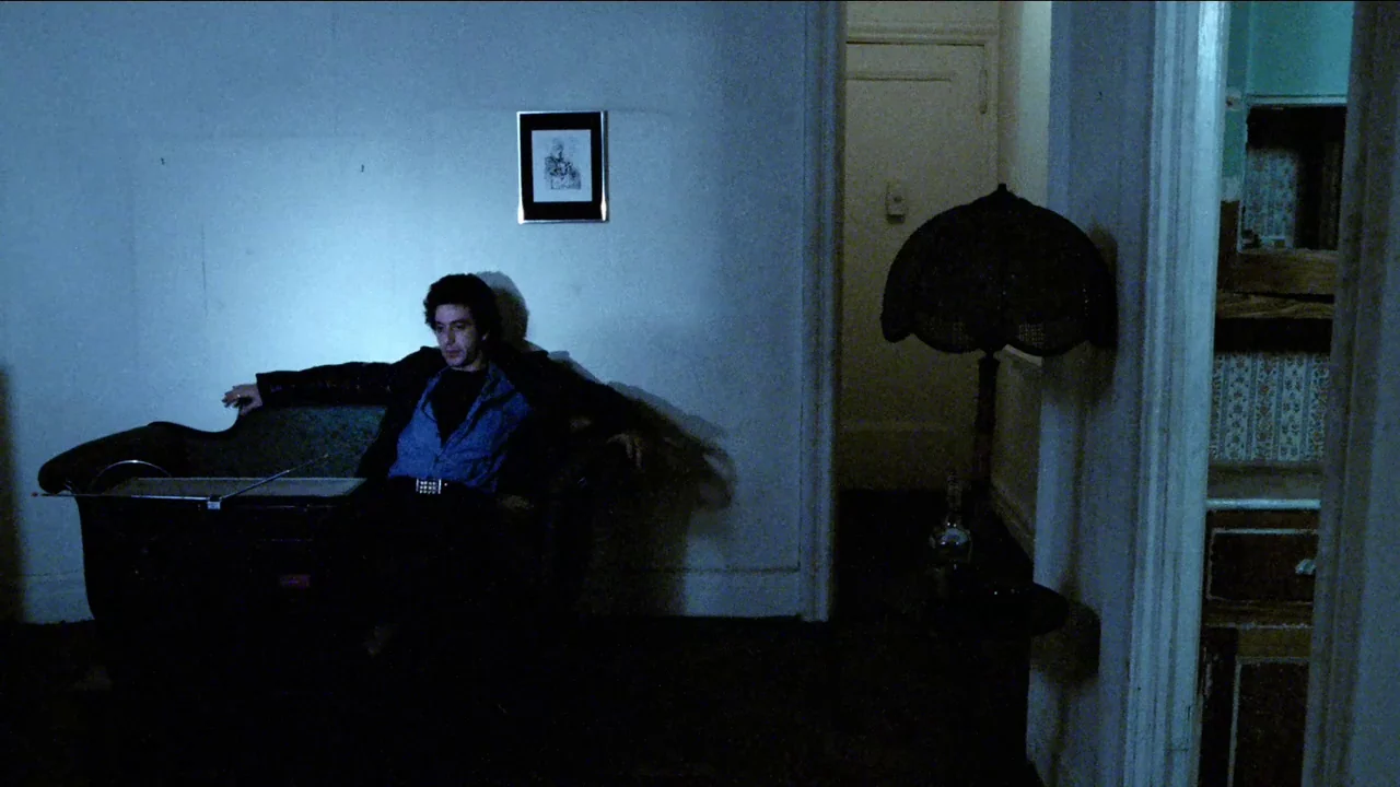 Cruising (1980) movie still: medium — A sparsely furnished apartment room with white walls and a dark sofa. A man sits alone on the sofa in dim…