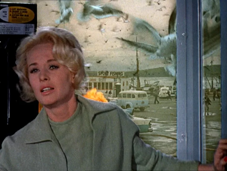The Birds movie still: medium — A woman stands inside a glass phone booth overlooking a busy waterfront harbor area. She looks toward the window as…