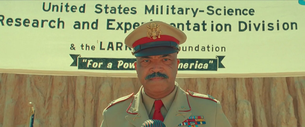Asteroid City (2023) movie still: medium — A man in military dress uniform stands in front of a white research sign. The background displays desert…
