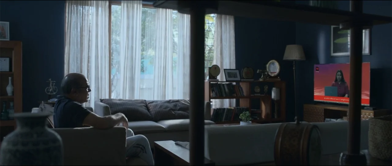 Garudan (2024) movie still: medium — A modern living room contains a grey couch, bookshelves, and a large television display. A middle-aged man with…