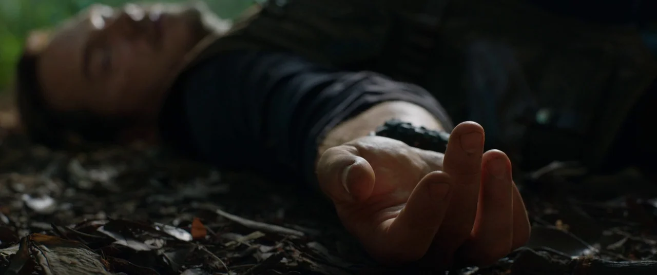 Jurassic World: Fallen Kingdom (2018) movie still: closeup — A man lies on a bed of dry brown leaves in a forest. His body is reclined while one…