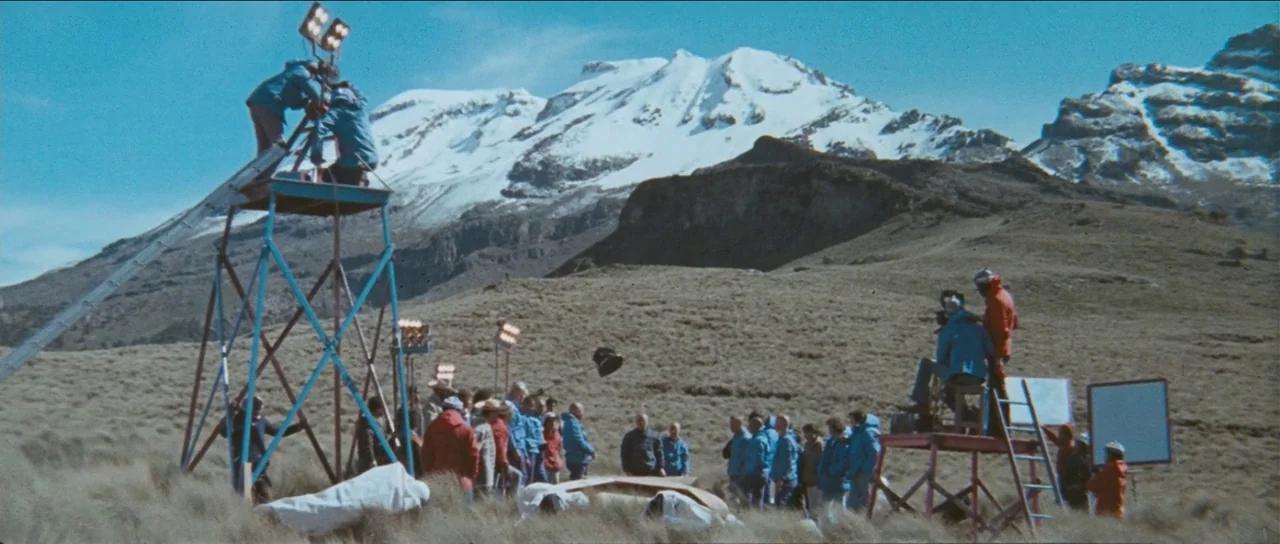 The Holy Mountain (1973) movie still: wide — A film crew works on a grassy mountainside with a large snow-capped peak behind. Men stand on metal…