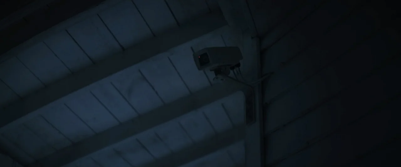 Wolf Man (2025) movie still: closeup — A dark, wooden building corner is visible under dim moonlight at night. An active security camera with a…