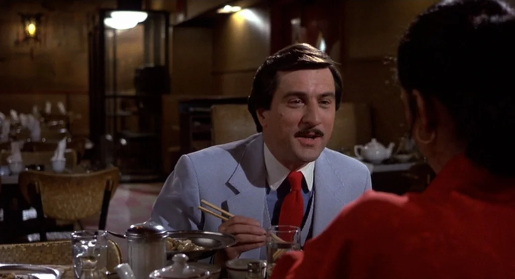 The King Of Comedy (1982) movie still: medium — An interior shot of a low-light dining establishment with wooden fixtures and dim lighting. A man in…