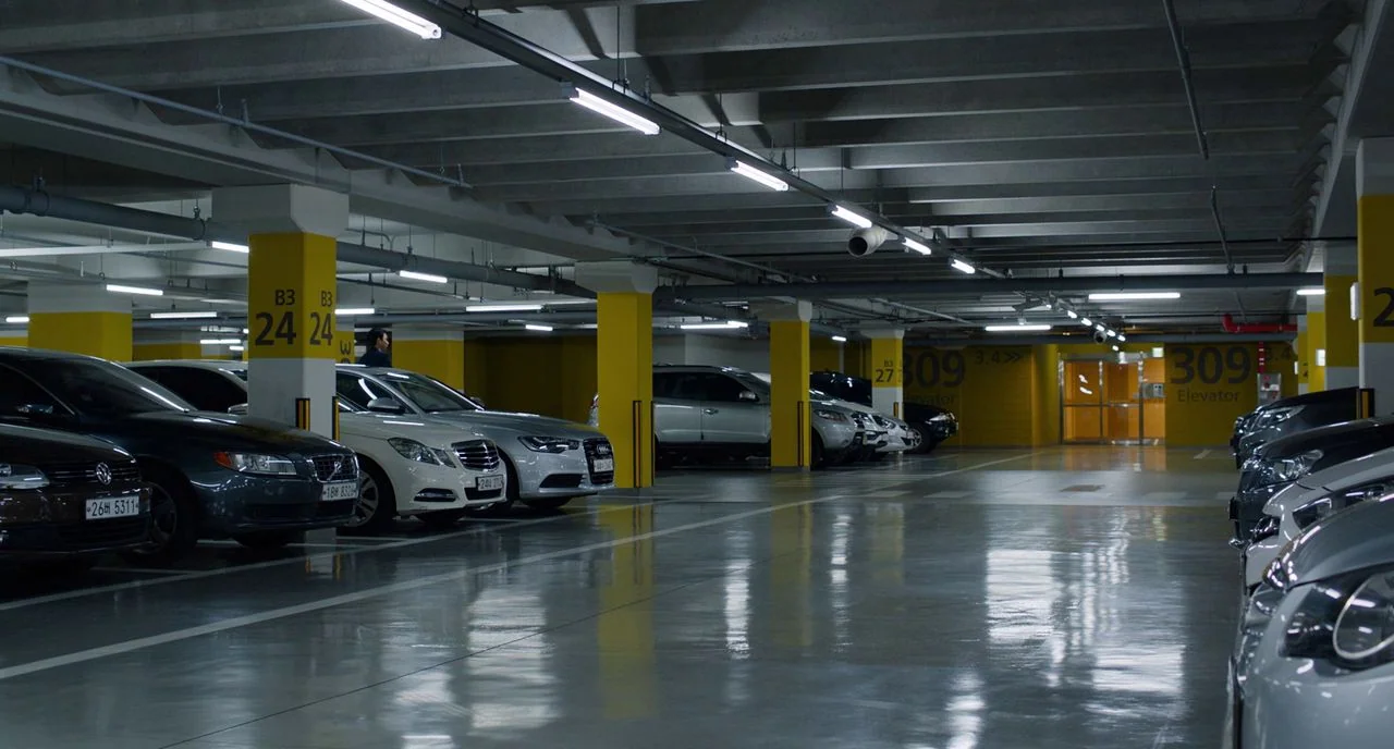 Train To Busan movie still: wide — A modern underground parking garage with bright yellow concrete columns and glossy grey floors. Multiple luxury…