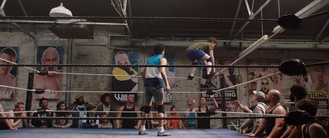 Fighting With My Family (2019) movie still: wide — An indoor wrestling ring with brick walls adorned with various professional wrestling promotional…