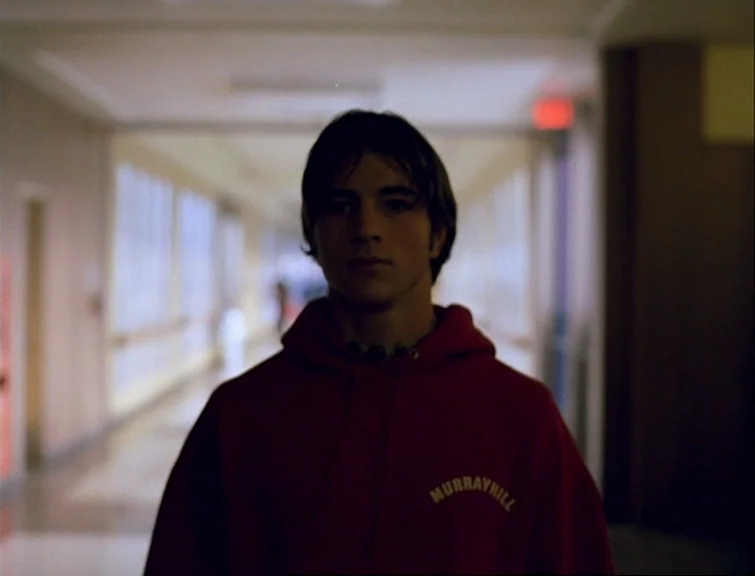 Elephant (2003) movie still: medium — A long school corridor with lockers and windows along the side wall in blurred focus. A young male stands…