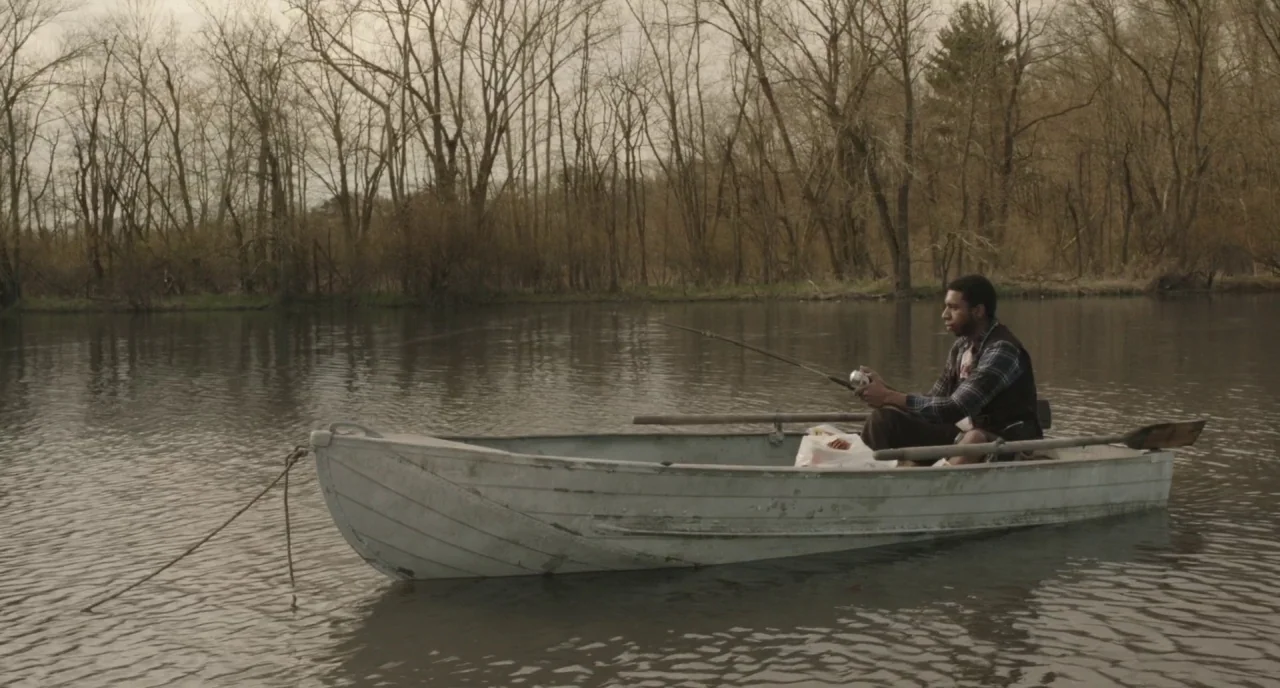 The Alchemist Cookbook (2016) movie still: wide — A man fishes from a weathered white rowboat on a calm, overcast lake. Bare deciduous trees line…
