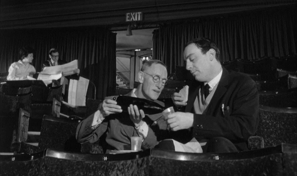A Hard Days Night (1964) movie still: medium — A dimly lit movie auditorium features rows of dark, fabric-covered seats and an exit sign. Two men…
