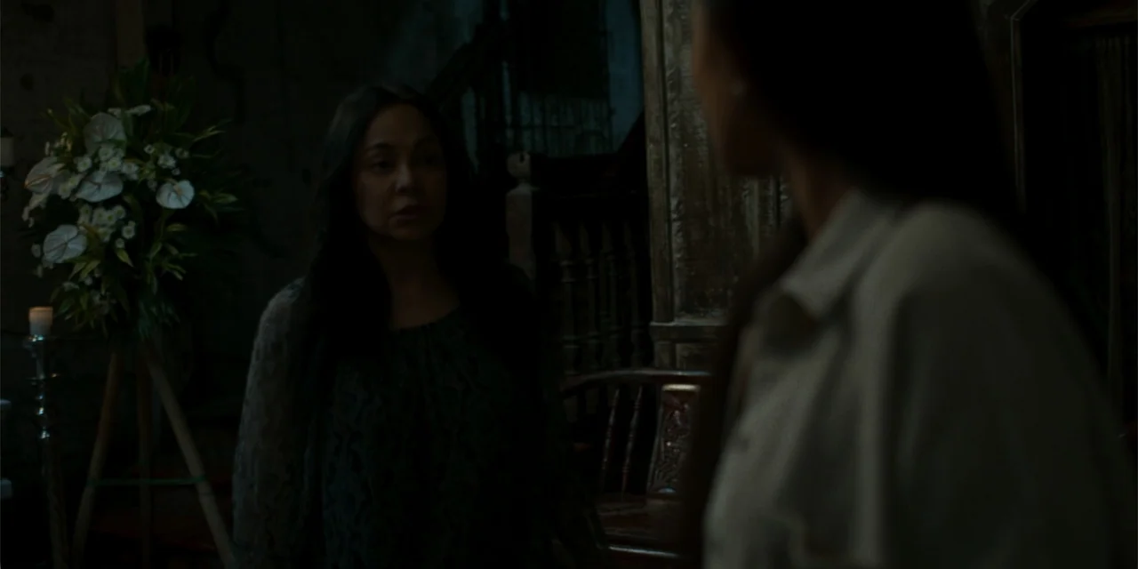 Nokturno (2024) movie still: medium — A dimly lit room contains a large flower arrangement on a wooden stand. Two women engage in a quiet dialogue…