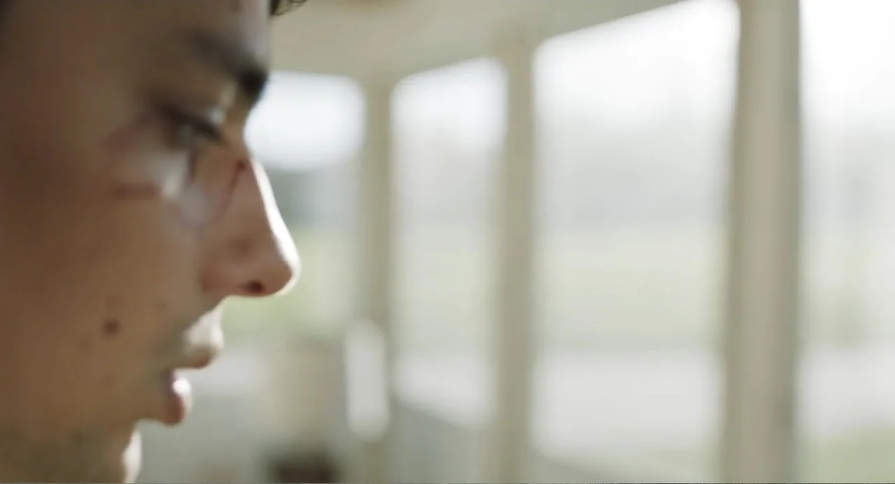 Salvation (2025) movie still: closeup — A bright sunroom features large glass panels that allow abundant natural light inside. A young man stands in…