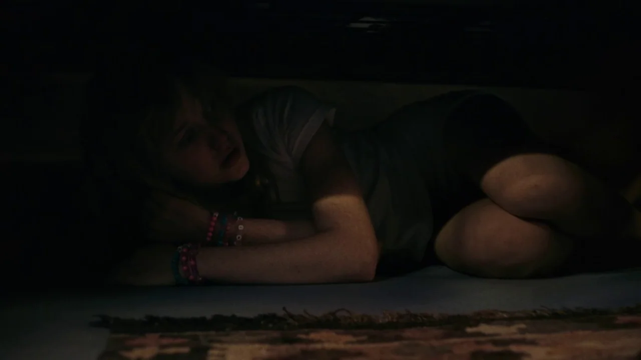 My First Summer (2020) movie still: medium — A teenager hides in the dark space underneath a bed frame. The floor is covered by a small patterned rug.