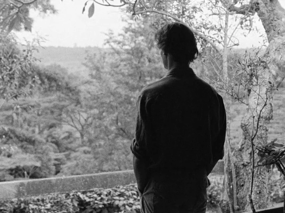 Tabu (2012) movie still: medium — An outdoor veranda overlooks a dense forest canopy under a bright overcast sky. A man stands at the edge with his…
