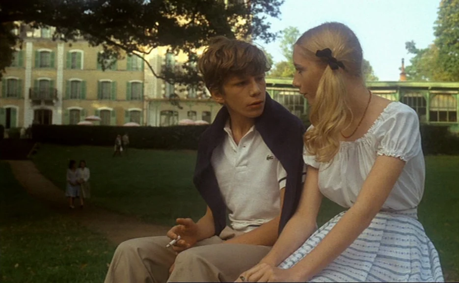 Murmur of the Heart (1971) movie still: medium — A teenage couple sits on a grassy lawn in front of a large manor house. The building has multiple…