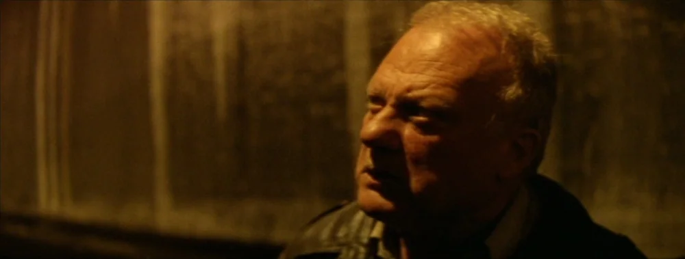I Stand Alone (Seul Contre Tous) (1998) movie still: closeup — An older man stands inside a dimly lit concrete structure with muted amber tones. He…