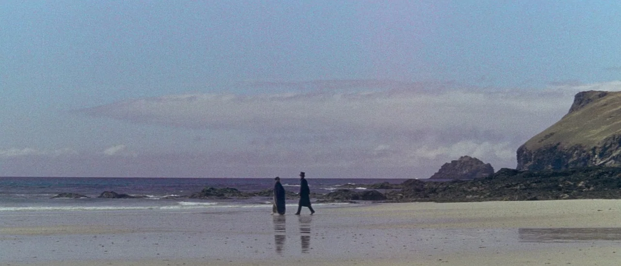 The Tomb of Ligeia (1964) movie still: wide — A vast, quiet beach stretches toward a distant rocky headland under a pale sky. Two people dressed in…
