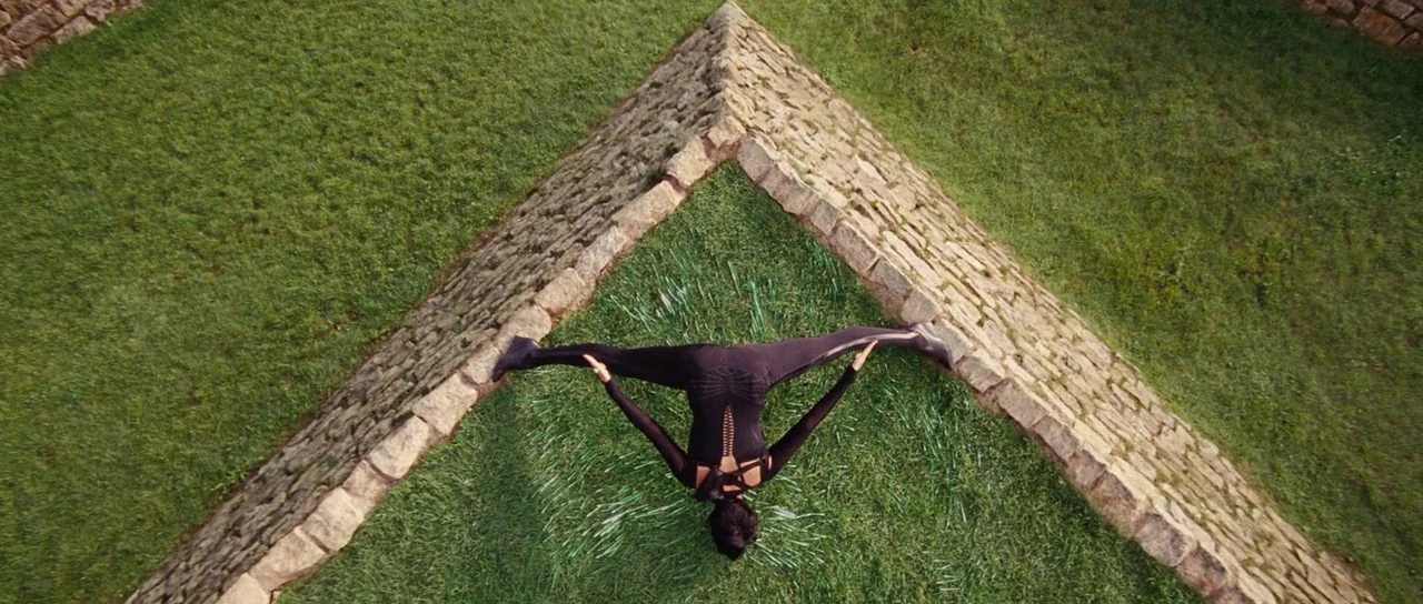 Aeon Flux (2005) movie still: wide — An outdoor area features grass enclosed by two converging weathered stone walls. A person wearing a black…