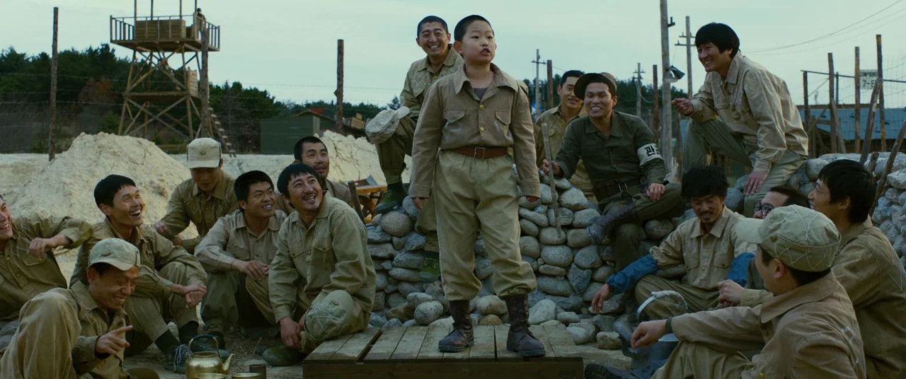 Swing Kids (2018) movie still: wide — A military encampment area features a wooden guard tower, barbed wire, and rock barriers. A young child…