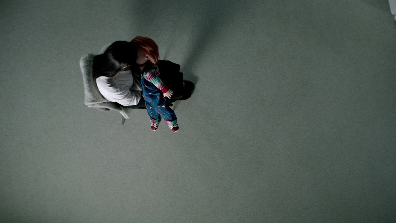 Cult of Chucky (2017) movie still: wide — An empty room with a plain carpet floor viewed from directly above. A woman sits on a chair while holding…