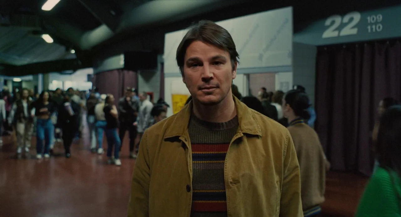 Trap (2024) movie still: medium — A crowded public concourse features signage labeled with the number 22. A man in a tan jacket stands centrally…