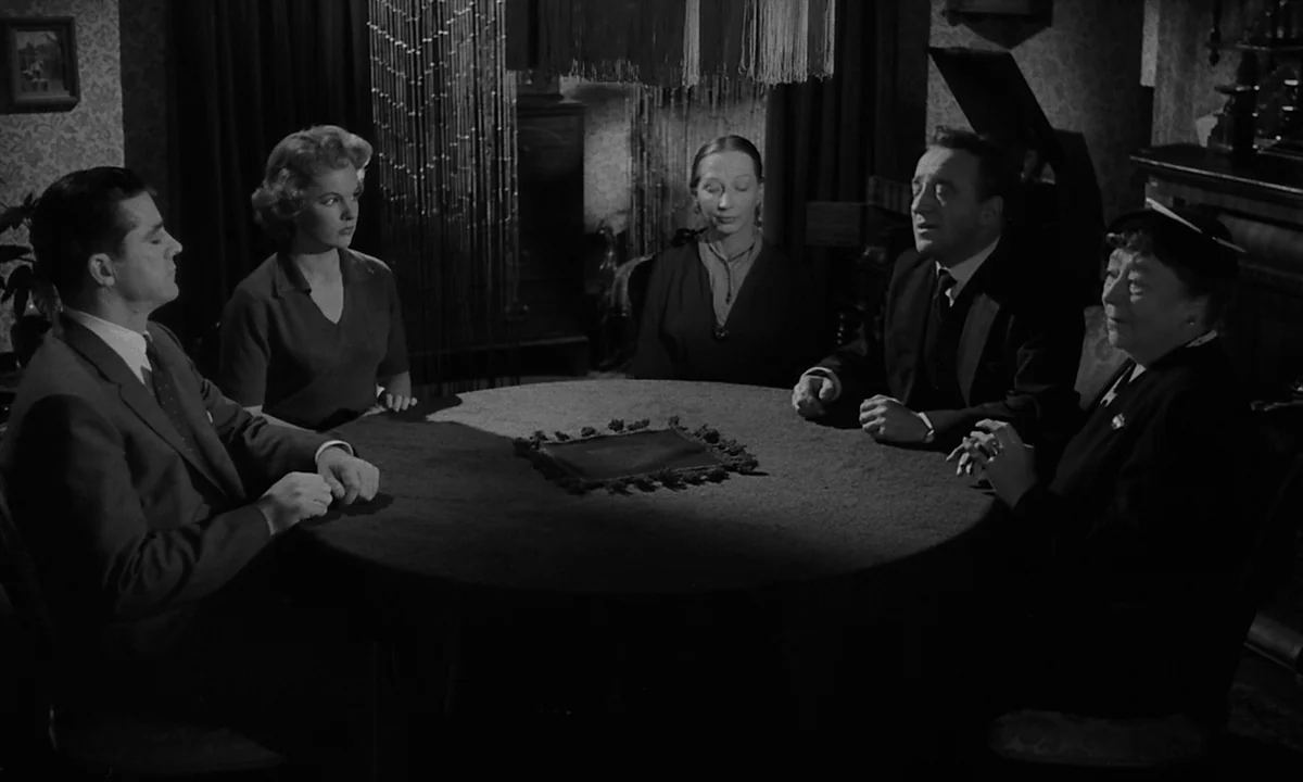 Night Of The Demon (1957) movie still: medium — A dimly lit room containing a circular table surrounded by five people in formal attire. The group…