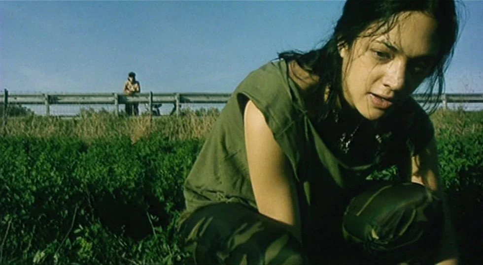 Scarlet Diva (2000) movie still: medium — A field of thick green vegetation stretches beneath a clear blue sky. A woman in camouflage gear crouches…