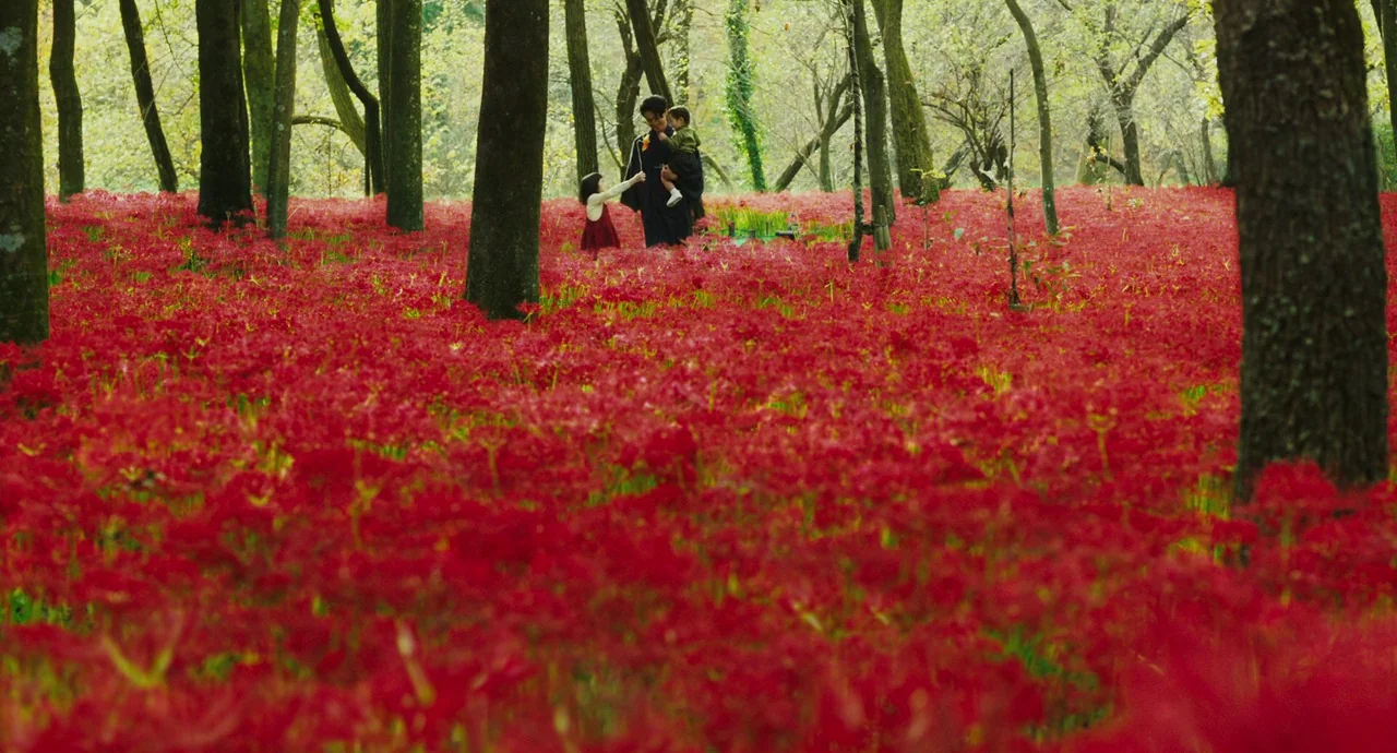 No Longer Human (2019) movie still: wide — A dense grove of trees surrounds a vast field of blooming red spider lilies. A woman holds one child and…