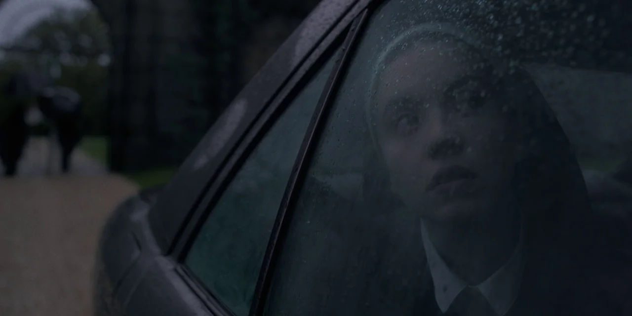 Immaculate (2024) movie still: medium — A person sits inside a vehicle with rain on the window glass. Outside the car, blurry figures holding…