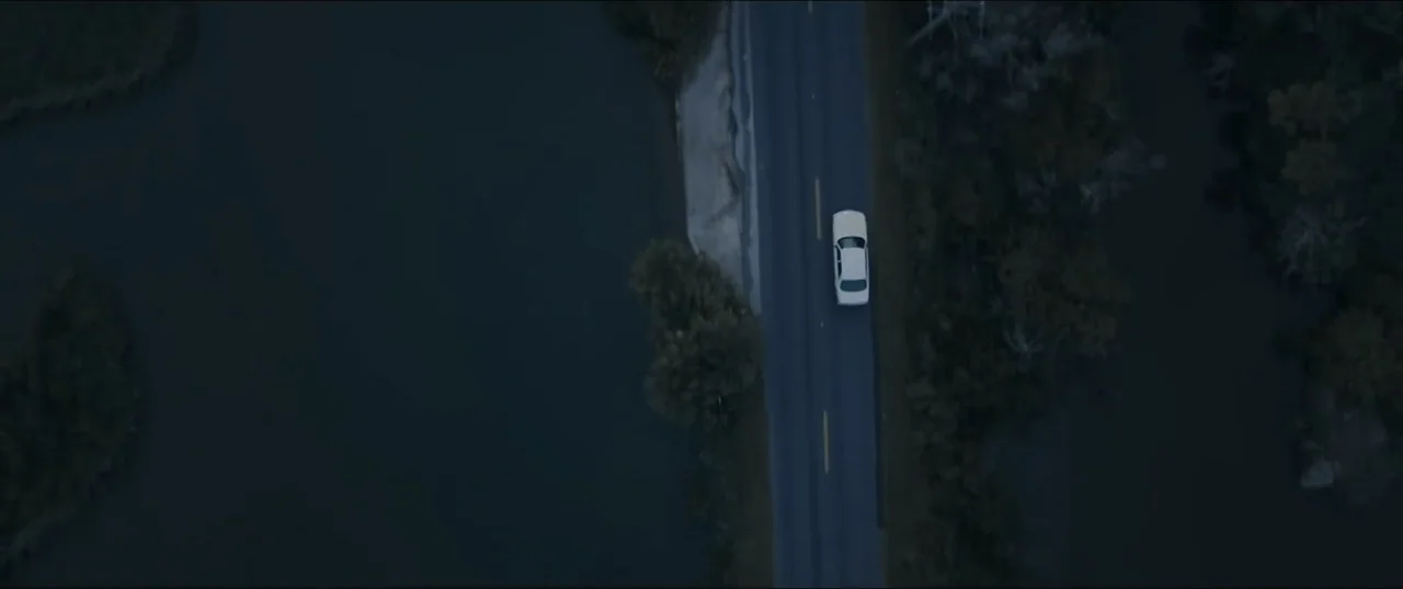 Offseason (2022) movie still: establishing — A straight asphalt road flanked by dark water and trees stretches through a landscape. A single white…