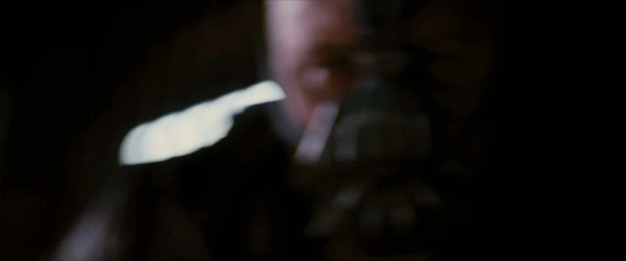 The Dark Knight Rises (2012) movie still: closeup — A figure is obscured by heavy darkness with only a sliver of light visible. The subject wears a…