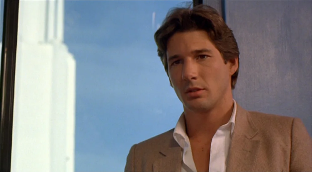 American Gigolo (1980) movie still: medium — An interior office space with a large window overlooking a clear blue sky. A man stands in front of the…