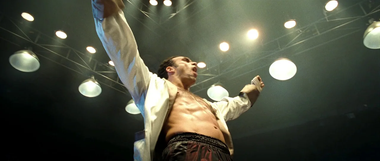 Ip Man 2 (2010) movie still: medium — A dimly lit arena interior with a dark background and numerous bright overhead lights. A man with his arms…