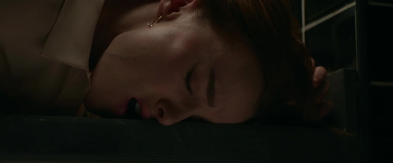Fair Play (2023) movie still: closeup — A person rests their head on a dark stone counter in a dark room. The subject appears unconscious or…
