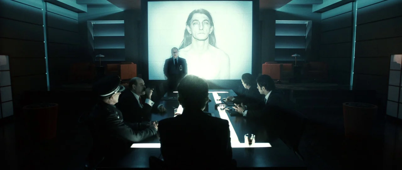 Daybreakers (2009) movie still: wide — A dark, modern conference room features a large projection screen displaying a man's mugshot. Five men in…