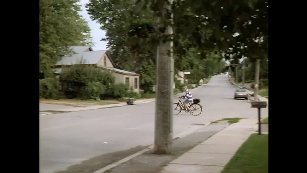 The Awakening (1995) movie still: wide — A quiet suburban street features mature trees, old houses, and a concrete utility pole. A young man rides a…