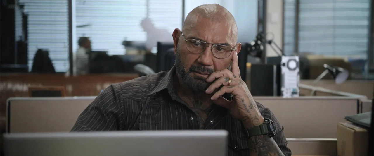 Trap House (2025) movie still: medium — A man works at a laptop inside a brightly lit corporate workspace filled with cubicle dividers. He looks…