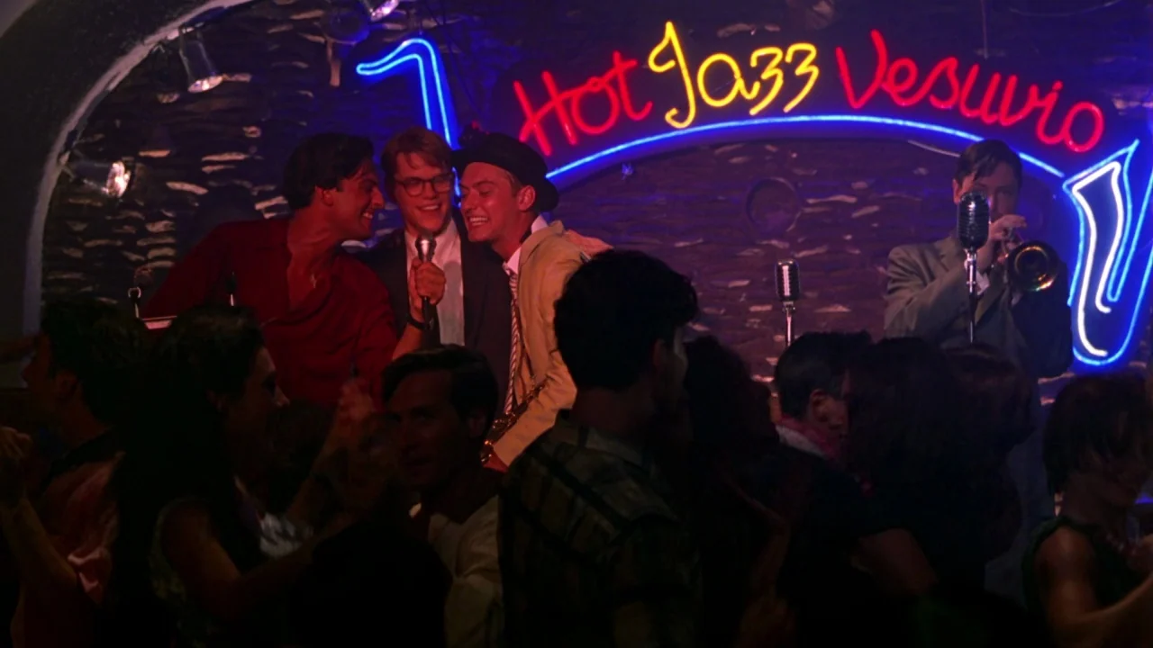 The Talented Mr. Ripley (1999) movie still: medium — A dimly lit club features a stone wall backdrop with a large neon sign. Several men perform…