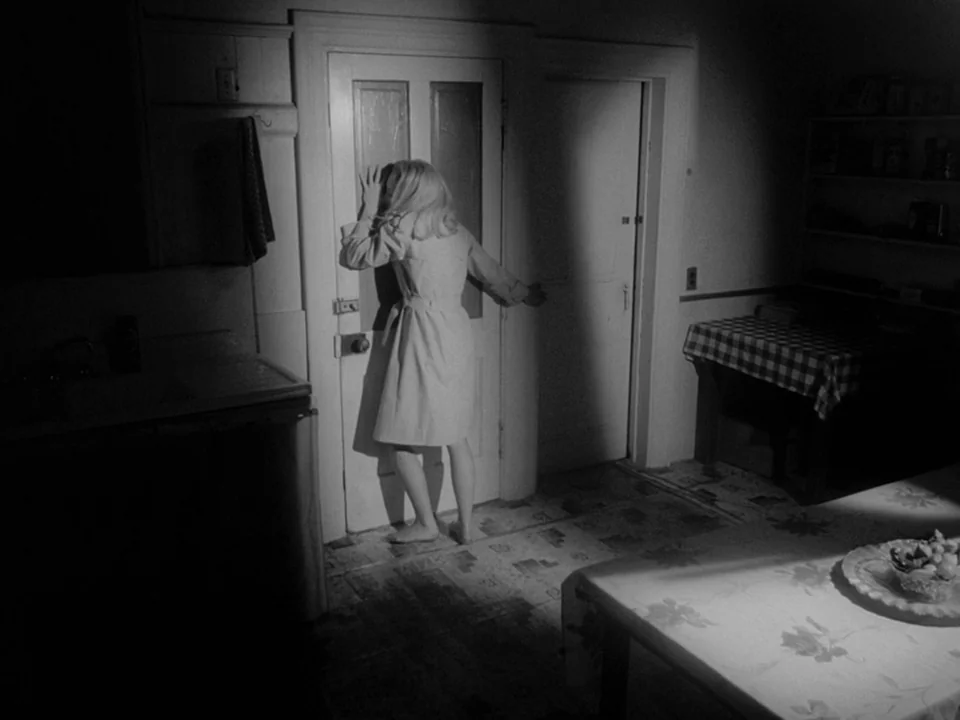 Night of the Living Dead (1968) movie still: fullbody — A dark, minimalist room featuring a wooden door and a small dining table. A young woman…