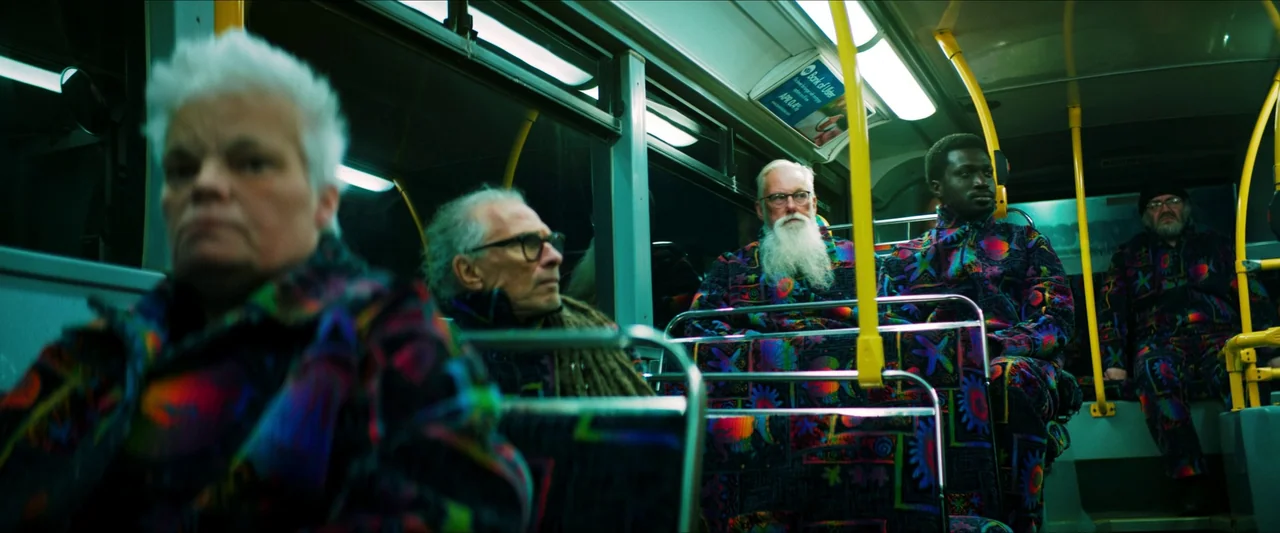 Kneecap (2024) movie still: medium — Interior of a city bus at night with passengers sitting in uniform rows. Overhead fluorescent lights illuminate…