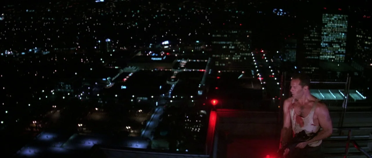 Die Hard (1988) movie still: wide — A high-angle view looking down from a rooftop overlooking a dense urban sprawl. A wounded man stands on the…