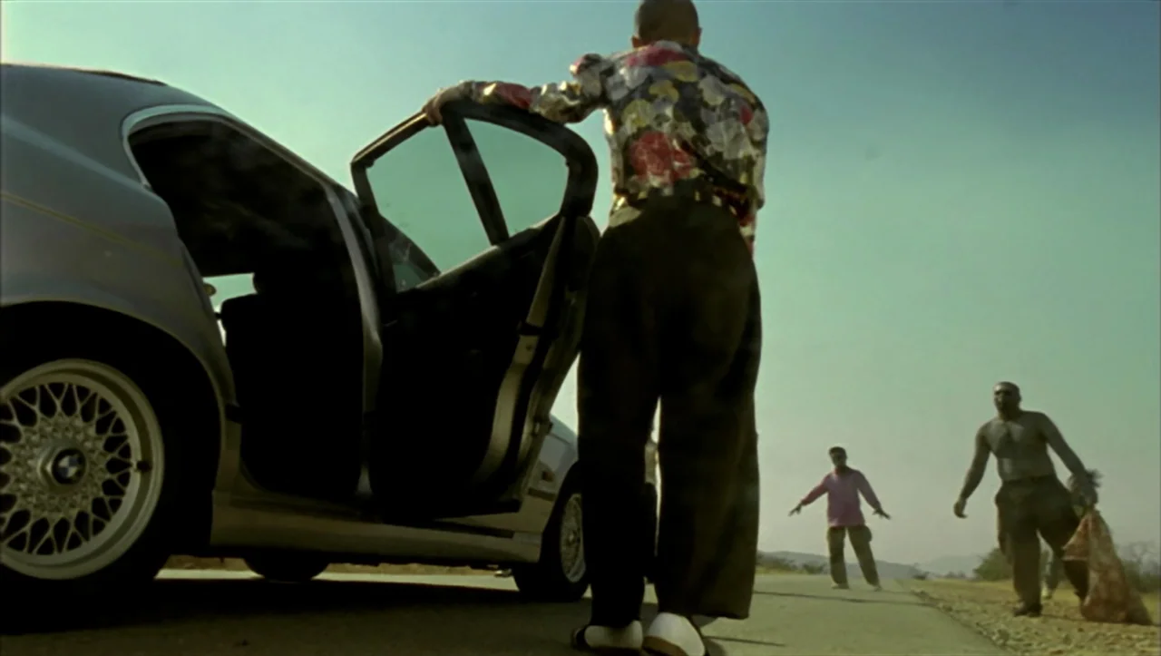 Wild Zero (1999) movie still: fullbody — A silver BMW is parked on a paved highway under a clear sky. A man stands at the car door as two other men…
