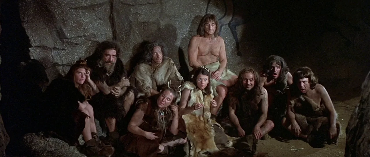 History of the World: Part 1 (1981) movie still: wide — A group of people wearing animal furs sit in a dimly lit rocky cave. They are huddled…