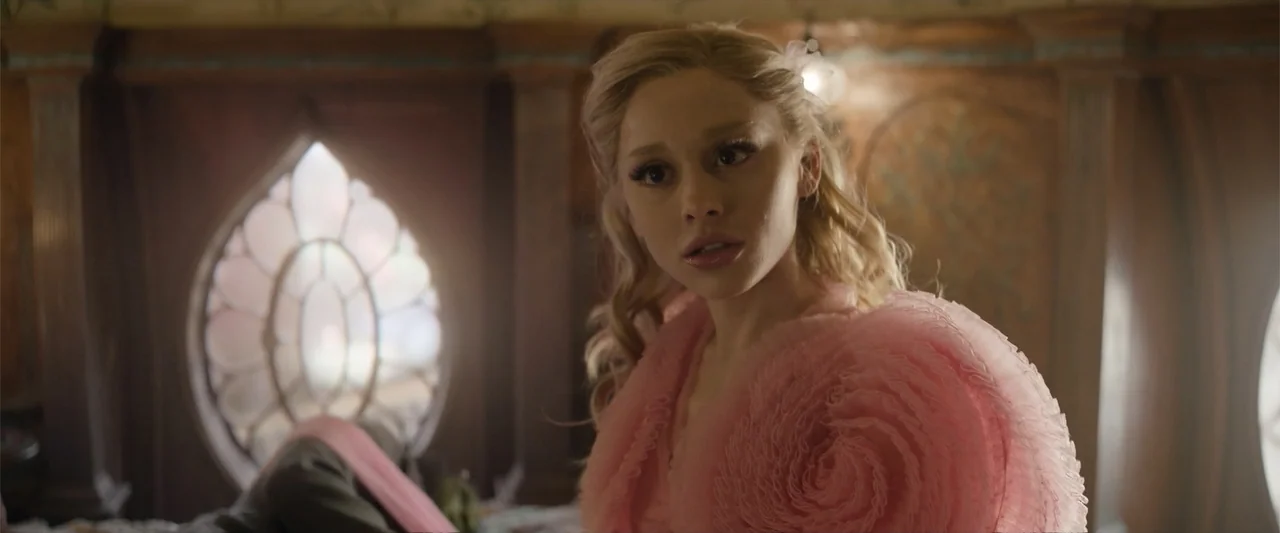 Wicked (2024) movie still: medium — An interior bedroom features wooden paneled walls and a distinctive arched stained glass window. A young woman…