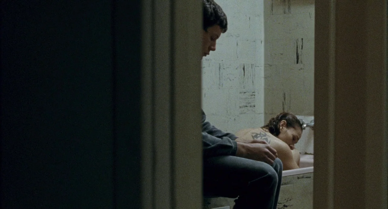 Snowtown (2011) movie still: medium — A cramped bathroom with light-colored, patterned wall tiles and a small bathtub. A young man sits nearby while…