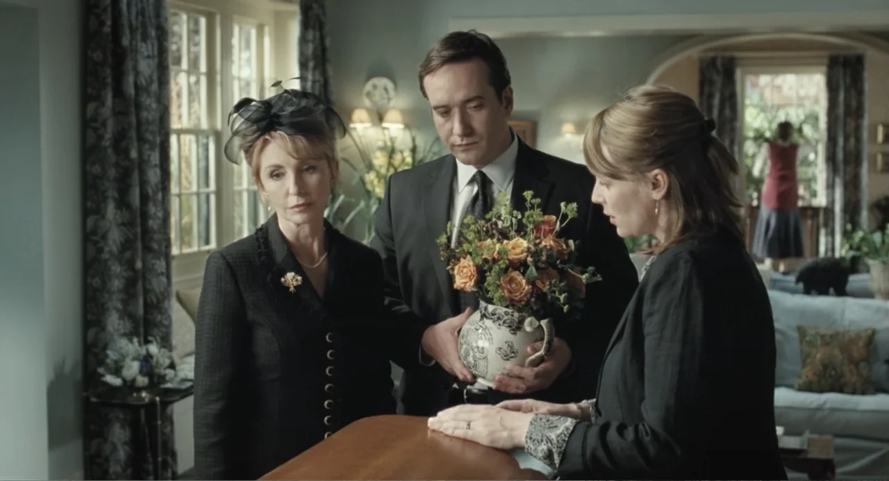 Death at a Funeral (2010) movie still: medium — A well-lit interior room contains a polished wooden casket surrounded by three grieving people. The…
